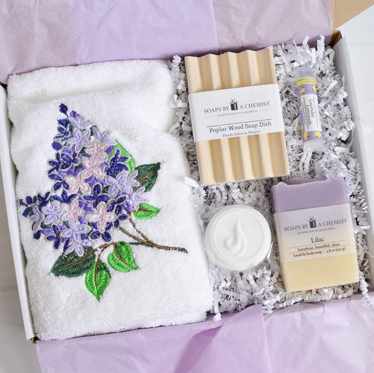 purple lilac floral gift set with lilac handmade soap, shower steamer, lilac embroidered hand towel, poplar wood soap dish, lavender lemonade lip balm in a gift box with purple tissue paper and white crinkle paper 
