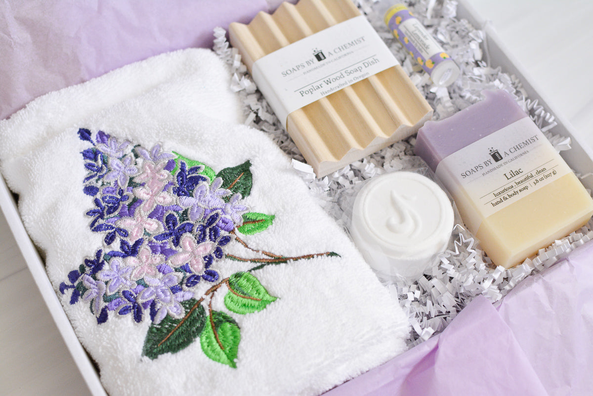 purple lilac floral gift set with lilac handmade soap, shower steamer, lilac embroidered hand towel, poplar wood soap dish, lavender lemonade lip balm in a gift box with purple tissue paper and white crinkle paper. side view