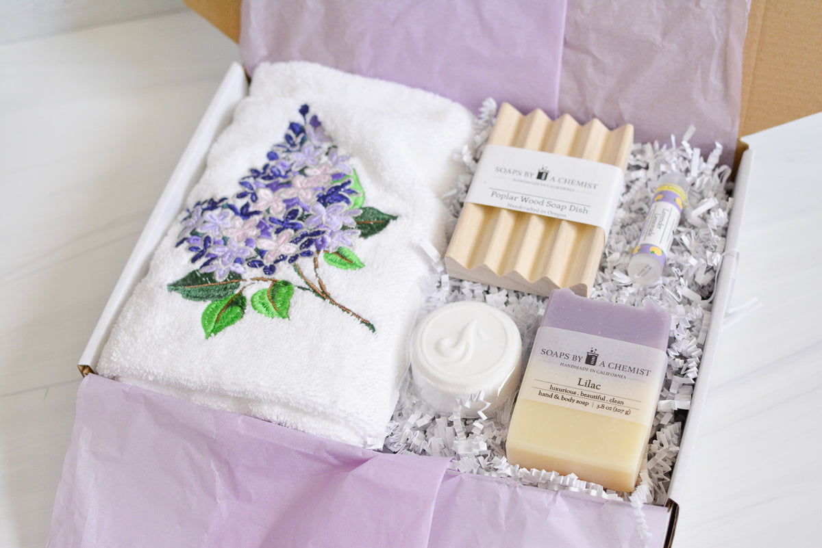purple lilac floral gift set with lilac handmade soap, shower steamer, lilac embroidered hand towel, poplar wood soap dish, lavender lemonade lip balm in a gift box with purple tissue paper and white crinkle paper. top view